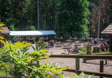 Eine idyllische Waldlichtung mit einem großen Zelt und vielen Sitzplätzen. Im Hintergrund sind Menschen und Fahrräder zu sehen, umgeben von Bäumen.