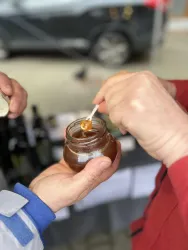 Ein Glas Honig wird mit einem Löffel entnommen. Es sind zwei Hände zu sehen, die an einem Verkaufsstand stehen.