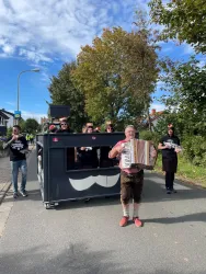 Eine Gruppe von Menschen in festlicher Kleidung zieht mit einem Wagen durch die Straße. Ein Mann spielt Akkordeon und sorgt für musikalische Unterhaltung.