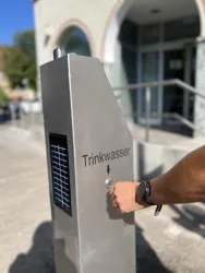 Ein moderner Trinkbrunnen aus Edelstahl mit der Aufschrift "Trinkwasser". Eine Hand betätigt den Wasserhahn.