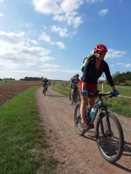 Eine Gruppe von Radfahrern fährt auf einem Schotterweg durch eine grüne Landschaft. Der Himmel ist blau mit einigen Wolken.