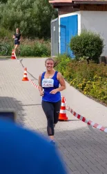 Eine Frau läuft fröhlich auf einem Wettkampf. Im Hintergrund sind einige Zuschauer und eine Laufstrecke mit Absperrung zu sehen.