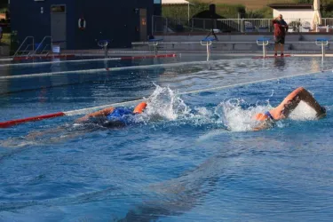 Zwei Schwimmer, die in einem blauen Schwimmbecken gegeneinander antreten. Die Sonne scheint und es spritzt Wasser.