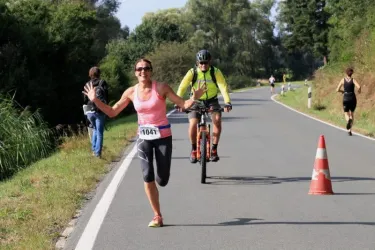 Eine fröhliche Läuferin in einem pinken Oberteil läuft auf einer Straße und winkt. Im Hintergrund sieht man einen Radfahrer und weitere Teilnehmer des Wettkampfs.