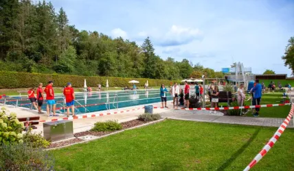 Ein Schwimmbad mit mehreren Personen, die sich auf dem Gelände aufhalten. Sicherheitspersonal in roten T-Shirts ist sichtbar, und das Poolareal ist abgesperrt.