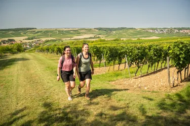 Zwei Frauen gehen lächelnd durch ein Weinberg. Im Hintergrund sind sanfte Hügel und eine klare blaue Himmel zu sehen.