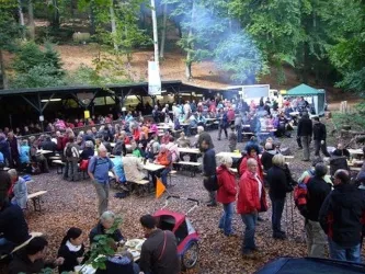 Eine große Menge von Menschen versammelt sich in einem Waldgebiet für ein Fest. Tische und Bänke sind aufgestellt, und es gibt Stände mit Essen und Getränken.