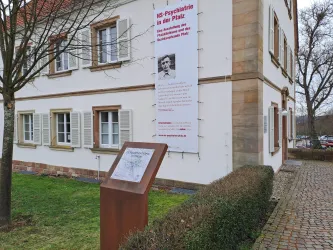 Ein historisches Gebäude mit einer Informationstafel im Vordergrund. An der Wand hängt ein großes Plakat mit Informationen zur Psychiatrie.