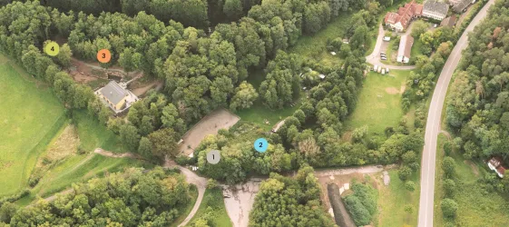 Eine Vogelperspektive eines Waldgebiets mit mehreren Wegen. Im Hintergrund sind Gebäude und eine Straße sichtbar.