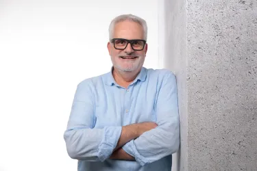 Ein lächelnder Mann mit Brille steht an einer Wand. Er trägt ein hellblaues Hemd und sieht freundlich aus.
