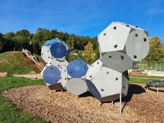 Ein moderner Spielplatz mit einer kletterfähigen Struktur in Form von geometrischen Formen. Im Hintergrund sind eine Rutsche und eine grüne Wiese zu sehen.