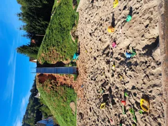 Ein Spielplatz mit einem Sandbereich und bunten Spielzeugen. Im Hintergrund sind grüne Hügel und ein klarer blauer Himmel zu sehen.