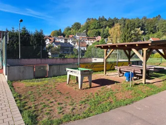 Ein Spielplatz mit einem Tischtennisplatte und einer überdachten Sitzgelegenheit. Im Hintergrund sind Häuser und Bäume zu sehen.