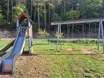 Ein Spielplatz mit einer Rutsche und einer Schaukel. Im Hintergrund sind Bäume und eine Wiese zu sehen.