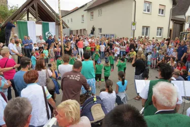 Eine lebhafte Versammlung von Menschen auf einer Straße bei einem festlichen Anlass. Kinder in grünen T-Shirts treten auf, während die Menge zuschaut und applaudiert.
