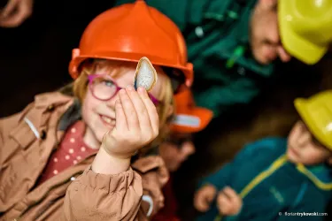 Ein fröhliches Kind mit einer Schutzhelm hält eine Muschel in der Hand. Im Hintergrund sind weitere Kinder und Erwachsene zu sehen, die ebenfalls Helme tragen.