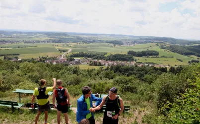 Eine Gruppe von Wanderern steht auf einem Hügel und genießt die Aussicht auf die grünen Felder und das Tal. Im Hintergrund sind weite Landschaften und ein kleines Dorf zu sehen.