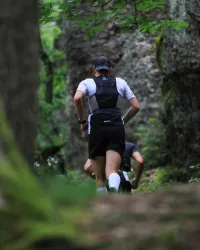 Ein Mann läuft auf einem schmalen Pfad durch den Wald. Im Hintergrund sind verschwommene Formen von Felsen und Bäumen zu sehen.