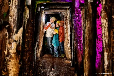 Eine Gruppe von Menschen erkundet einen dunklen Stollen mit Holzstützen. Sie tragen Helme und halten Taschenlampen, während farbige Lichter den Tunnel beleuchten.