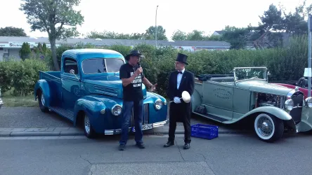 Zwei Oldtimer-Fahrzeuge stehen auf der Straße, ein blauer Pickup und ein weißes Auto. Ein Mann im Anzug spricht mit einem anderen Mann, der ein T-Shirt trägt.