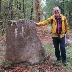 Ein Mann in gelber Jacke steht neben einem großen Stein in einem Wald. Der Stein trägt eine Inschrift und das Umfeld ist herbstlich mit bunten Blättern.