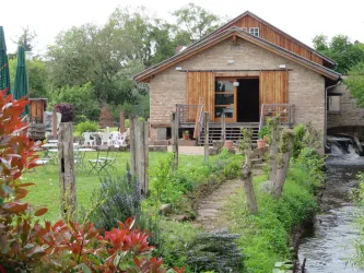 Ein rustikales Haus mit Holzverkleidung, umgeben von Grün und einem kleinen Wasserlauf. Auf der Wiese stehen Stühle und Tische zum Entspannen.