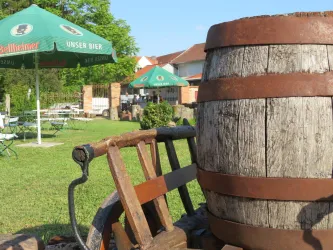 Ein Holzfass steht auf einem Gartenstuhl. Im Hintergrund sind grüne Wiesen und Schirme zu sehen.