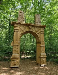Ein kunstvoll gestalteter Bogen steht im Wald zwischen Bäumen. Der Bogen hat zahlreiche Verzierungen und ist aus hellem Material gefertigt.