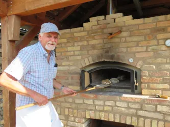 Ein älterer Mann steht in der Nähe eines gemauerten Ofens und hält eine Holzschaufel. Im Ofen sind mehrere Brote sichtbar, während er lächelnd in die Kamera schaut.