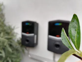 Zwei schwarze Geräte an einer Wand, umgeben von Pflanzen. Im Vordergrund ist ein Blatt mit Wassertropfen zu sehen.
