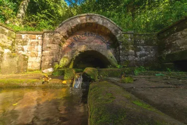 Ein alter Steinbogen, der einen Wasserlauf zeigt. Umgeben von üppigem Grün und Bäumen.