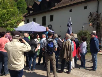 Eine Gruppe von Touristen versammelt sich um einen Führer in einer malerischen Umgebung. Im Hintergrund ist ein charmantes Gebäude und Bäume zu sehen.