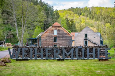 Ein rustikales Gebäude mit dem Schriftzug "WEIHERMÜHLE" im Vordergrund. Umgeben von Bäumen und einer grünen Landschaft.