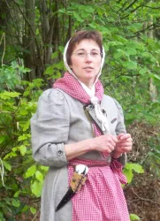Eine Frau in traditioneller Kleidung steht vor einem grünen Hintergrund. Sie trägt einen Kopftuch und hat einen freundlichen Ausdruck.