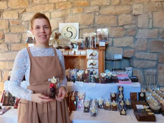 Eine Frau steht an einem Tisch mit verschiedenen Schokoladenprodukten. Im Hintergrund sind weitere Süßigkeiten und eine dekorative Wand aus Stein zu sehen.