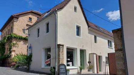 Ein helles und freundliches Gebäude in einer malerischen Straße. Der Himmel ist blau mit wenigen Wolken und es gibt grüne Pflanzen um das Haus herum.