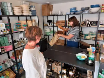 Eine Kundin in einem schicken Laden beobachtet eine Verkäuferin, die Waren einpackt. Regale sind mit verschiedenen Produkten und Utensilien gefüllt.