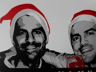 Zwei Männer lächeln und tragen Weihnachtsmann-Hüte. Der Hintergrund ist schlicht und einfarbig.
