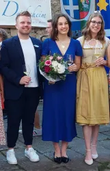 Drei Personen stehen zusammen, lächeln und halten Getränke in den Händen. Sie tragen elegante Kleidung und sind vor einer festlichen Kulisse zu sehen.