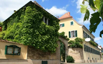 Ein charmantes Gebäude mit grüner Bewachsung und historischen Fenstern. Im Hintergrund sind weitere, hell gestrichene Gebäude und ein klarer Himmel zu sehen.