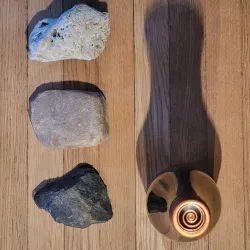 Drei Steine in verschiedenen Formen liegen auf einem Holzuntergrund. In der Mitte befindet sich ein goldener Gegenstand mit spiralförmigem Design.
