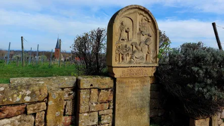 Ein historischer Stein mit Reliefs steht in einer ländlichen Umgebung. Im Hintergrund sind Weinberge und ein kirchturmähnliches Gebäude zu sehen.