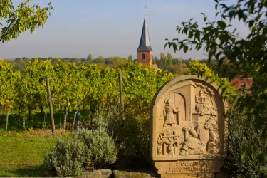 Eine steinerne Reliefskulptur steht inmitten von Weinreben. Im Hintergrund sieht man einen Kirchturm und eine ländliche Landschaft.