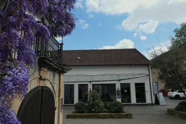 Ein historisches Gebäude mit modernen Fenstern und einem schönen Garten. Im Vordergrund blühen lila Pflanzen und der Himmel ist teilweise bewölkt.