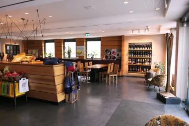 Ein modernes Café mit Holzinterieur und großen Fenstern. An der Wand stehen Regale mit Flaschen, während Tische und Stühle zum Verweilen einladen.