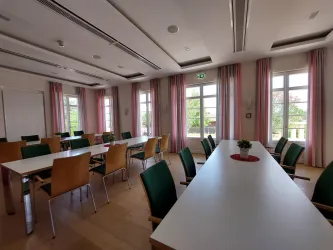 Ein moderner Besprechungsraum mit großen Fenstern und hellen Vorhängen. Tische und Stühle sind ordentlich angeordnet, und in der Mitte steht eine kleine Pflanze.