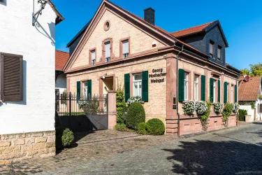 Ein charmantes Weingut mit einer roten Backsteinfassade und grünen Fensterläden. Die Umgebung ist gepflegt mit Büschen und einem ruhigen Straßenverlauf.