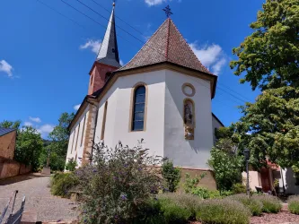 Eine kleine Kirche mit einem spitzen Turm und einem roten Dach. Umgeben von üppigem Grün und blühenden Pflanzen.