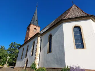 Eine Kirche mit einem hohen, spitzen Turm und einem schlichten weißen Äußeren. Der Himmel ist klar und blau, umgeben von Bäumen und Grünflächen.