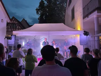 Ein Konzert im Freien mit einer Band unter einem Pavillon. Die Zuschauer stehen versammelt und genießen die Musik bei dämmerndem Licht.